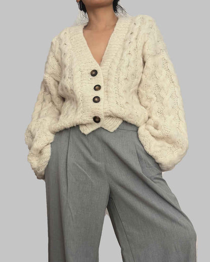 Person wearing a cream knitted cardigan and gray pants on a plain background