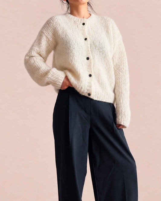 A model wearing a cream-colored, textured alpaca knit cardigan with black buttons and dark navy wide-leg trousers, standing against a solid pale pink studio backdrop.