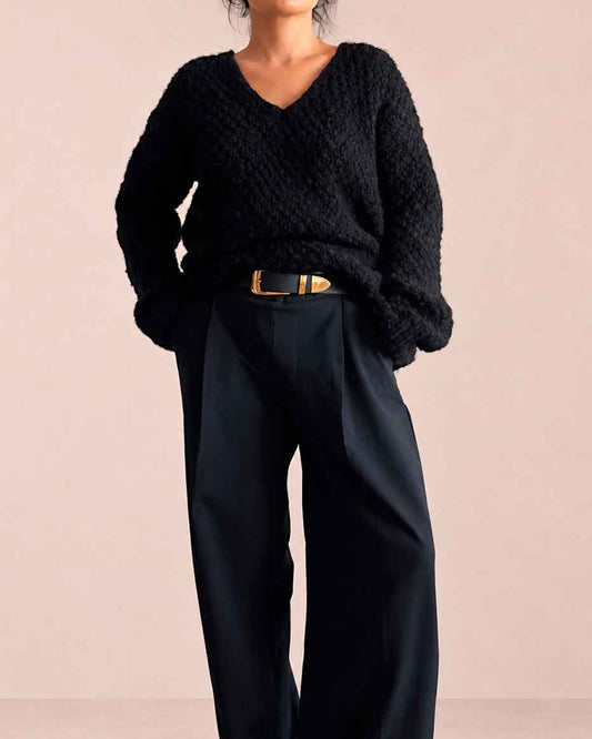 A model wearing a chunky black knit V-neck knit alpaca sweater and black wide-leg trousers with a gold belt, standing against a solid pale pink studio background.