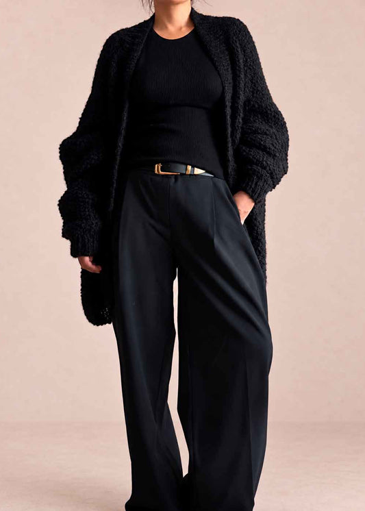 A model wearing a black textured open-front alpaca knit cardigan, a black ribbed top, and black wide-leg trousers with a gold-tone belt, set against a solid pale pink studio background.
