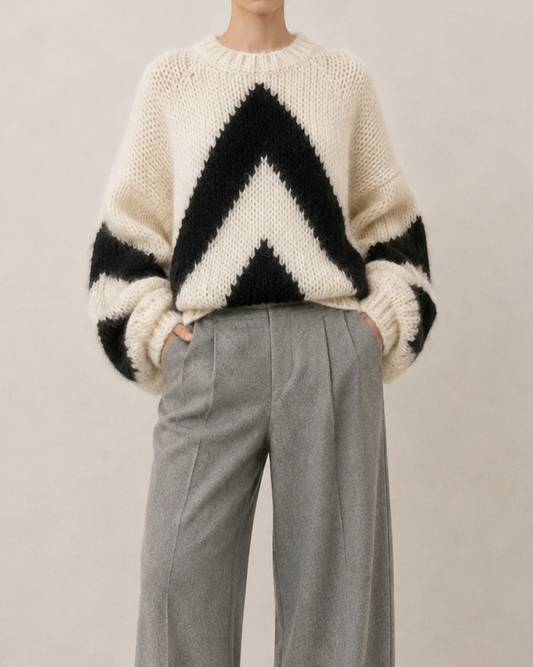 A model stands against a plain beige studio background wearing a cream and black oversized knit alpaca sweater with a bold chevron pattern and wide-leg gray trousers.